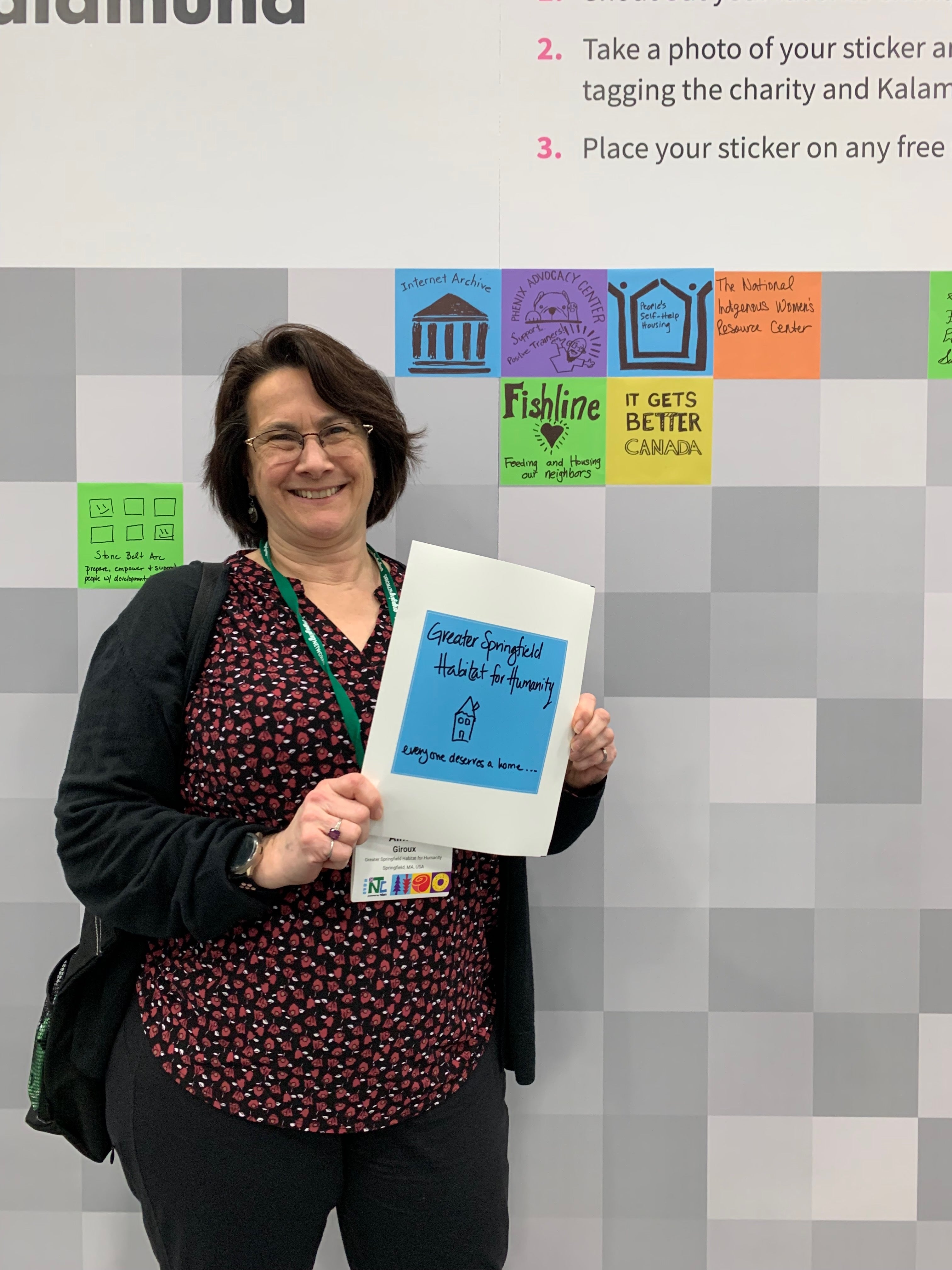 Aimee Giroux, a tall white woman grinning with short brown hair holding a sign that says 'Greater Springfield Habitat for Humanity' with a drawing of a house that states 'everyone deserves a home.'