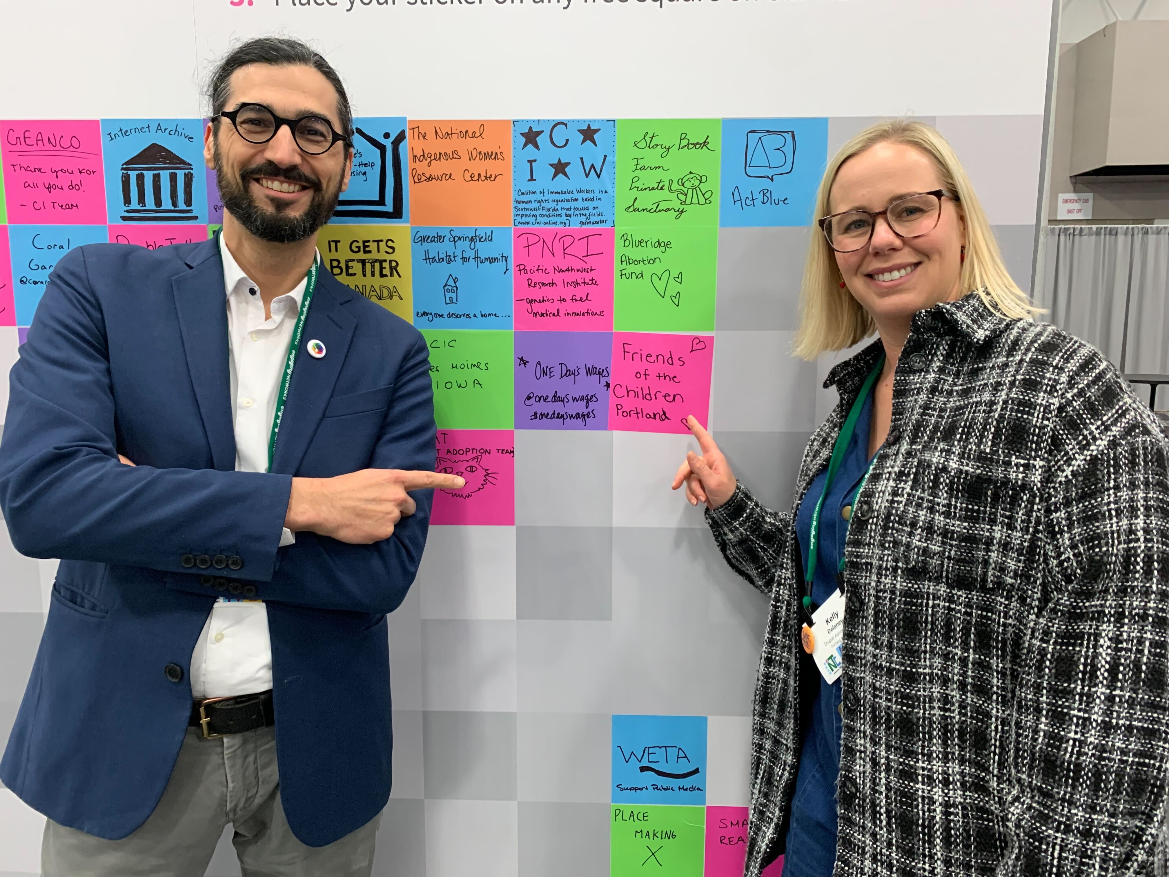 Andrew Mallis, tall Persian man with glasses and a beard with his hair tied back in a pony tail, posing with Kelly Delaney, white woman with blond hair and glasses and a plaid jacket, both pointing at the Kalamuna donation wall. 