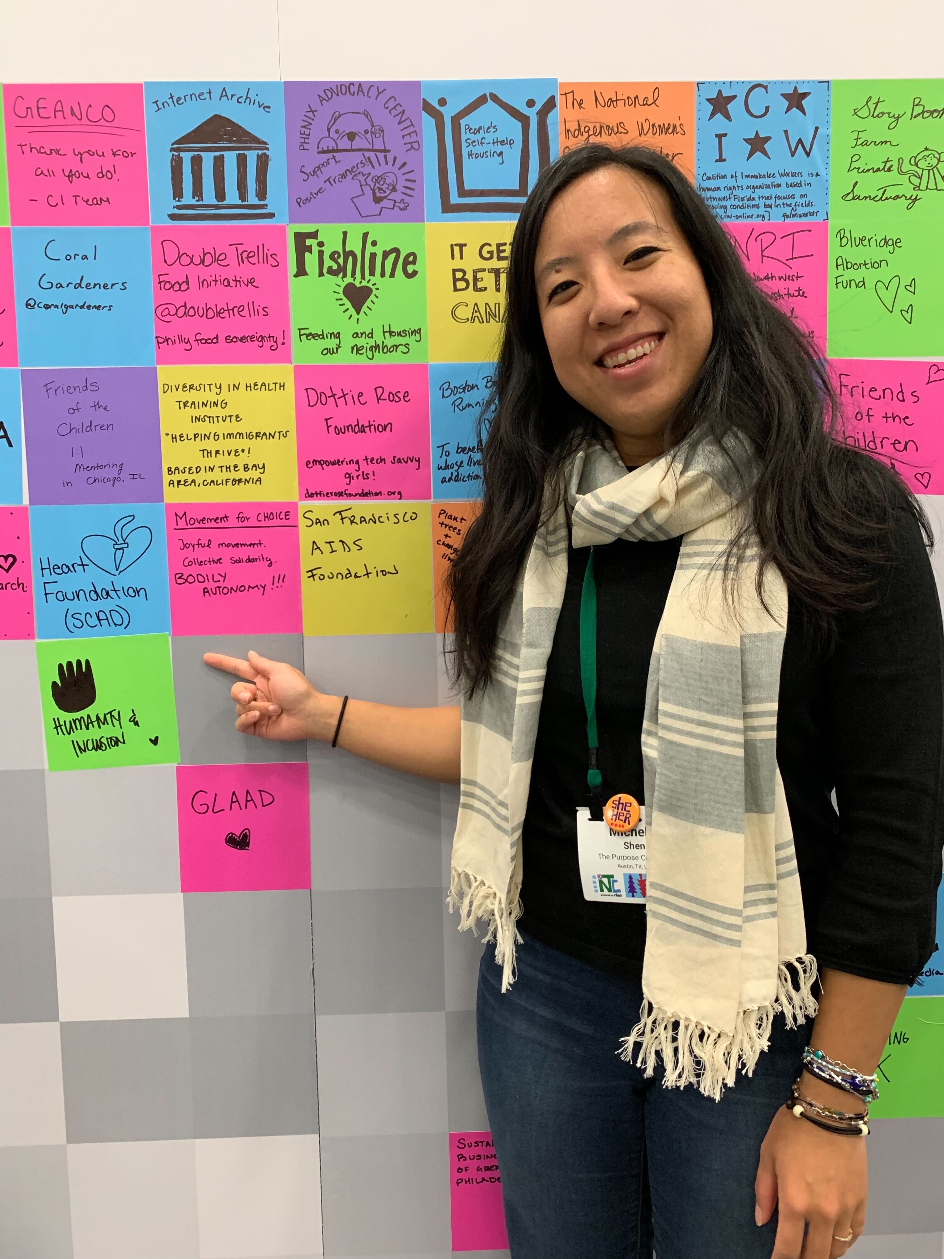 Michelle Shen, an Asian woman with long black hair wearing a white and grey striped scarf, pointing at the Kalamuna donation wall, with a colored in hand and the words 'Humanity and Inclusion' with a heart. 