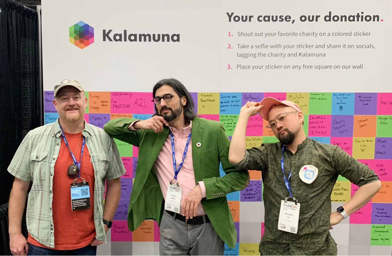 Bob, Andrew, and Ken stand in front of Kalamuna's booth wall. The wall is filled with colorful stickers that attendees have written names of charities on.