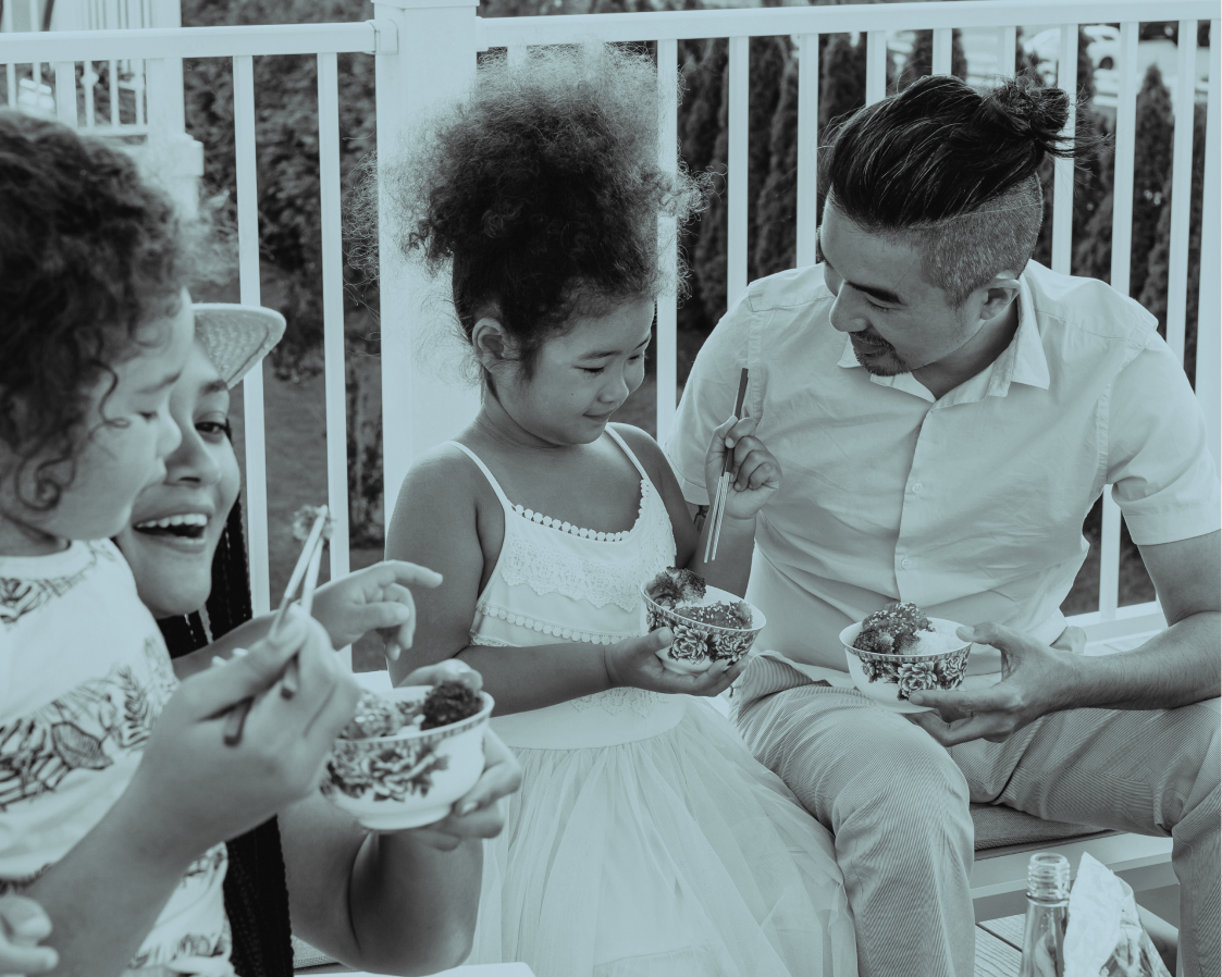 A family of 4 sits happily on a patio eating food with chopsticks.