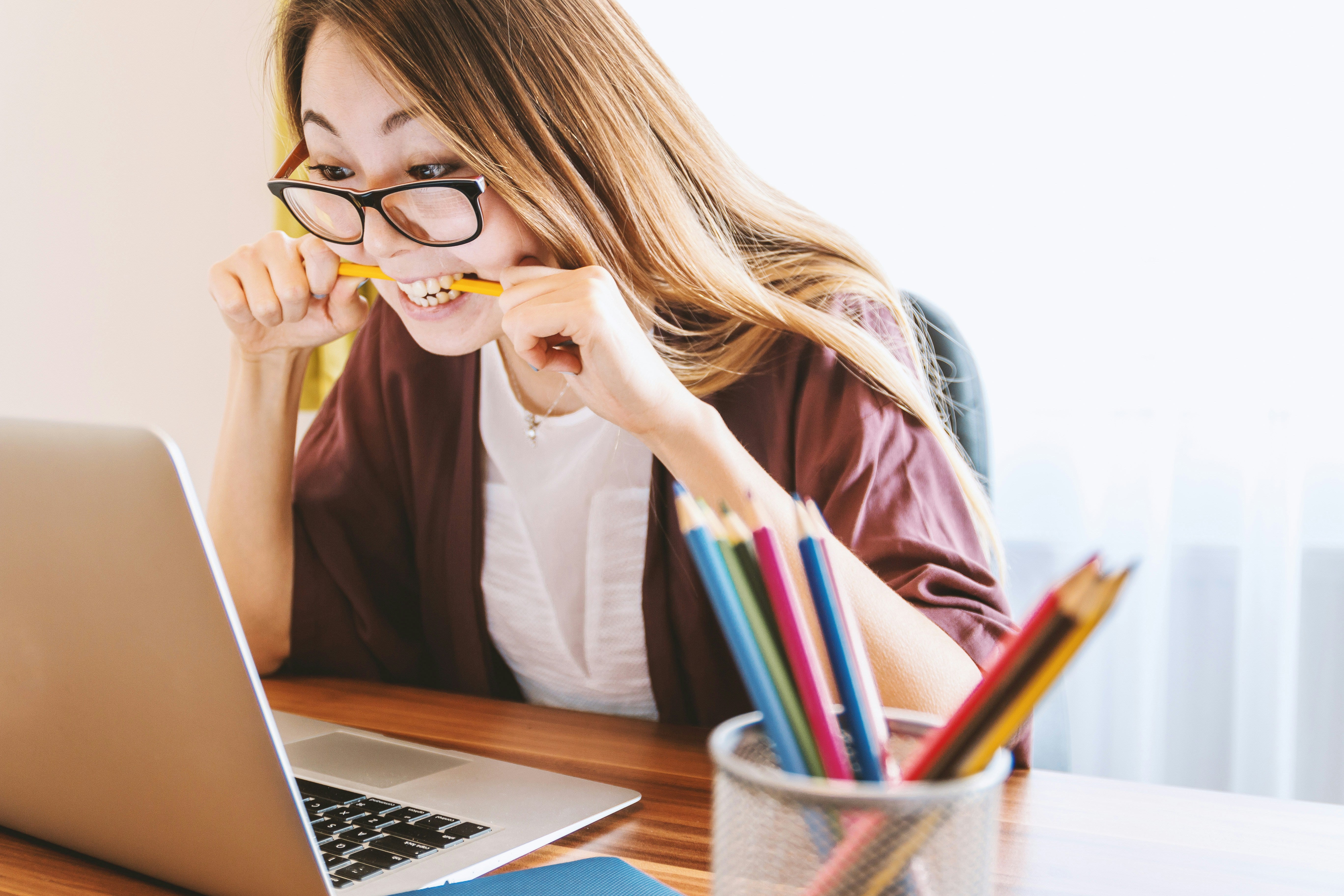 A young woman with glasses and long hair clenches a pencil between her teeth while looking at her laptop