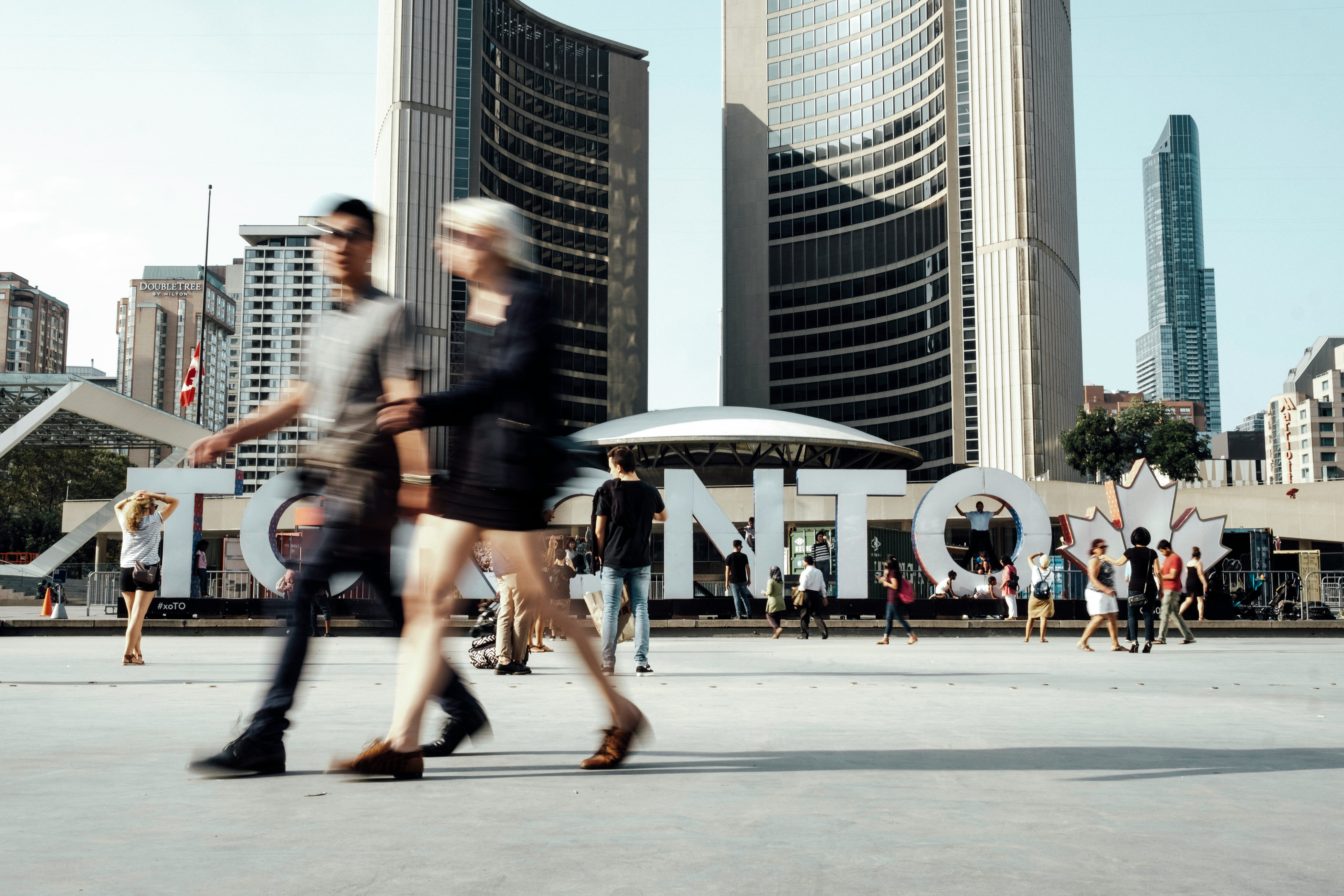 Pedestrians walk in motion blur past the Toronto sign at Nathan Phillips Square, with City Hall towers behind them.
