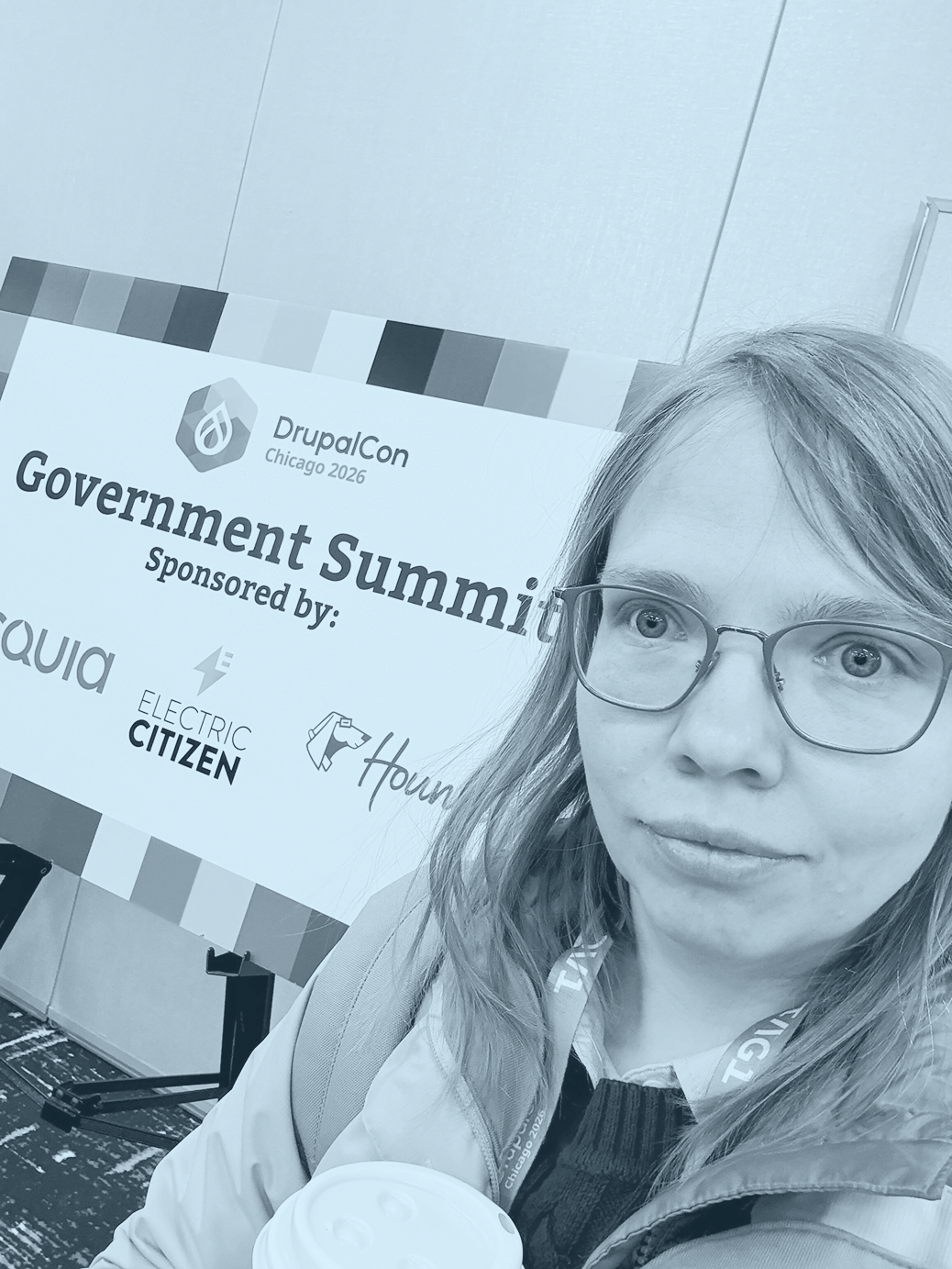 A selfie of Anya, white woman with glasses, standing in front of the DrupalCon Chicago Government Summit Banner holding a coffee cup.