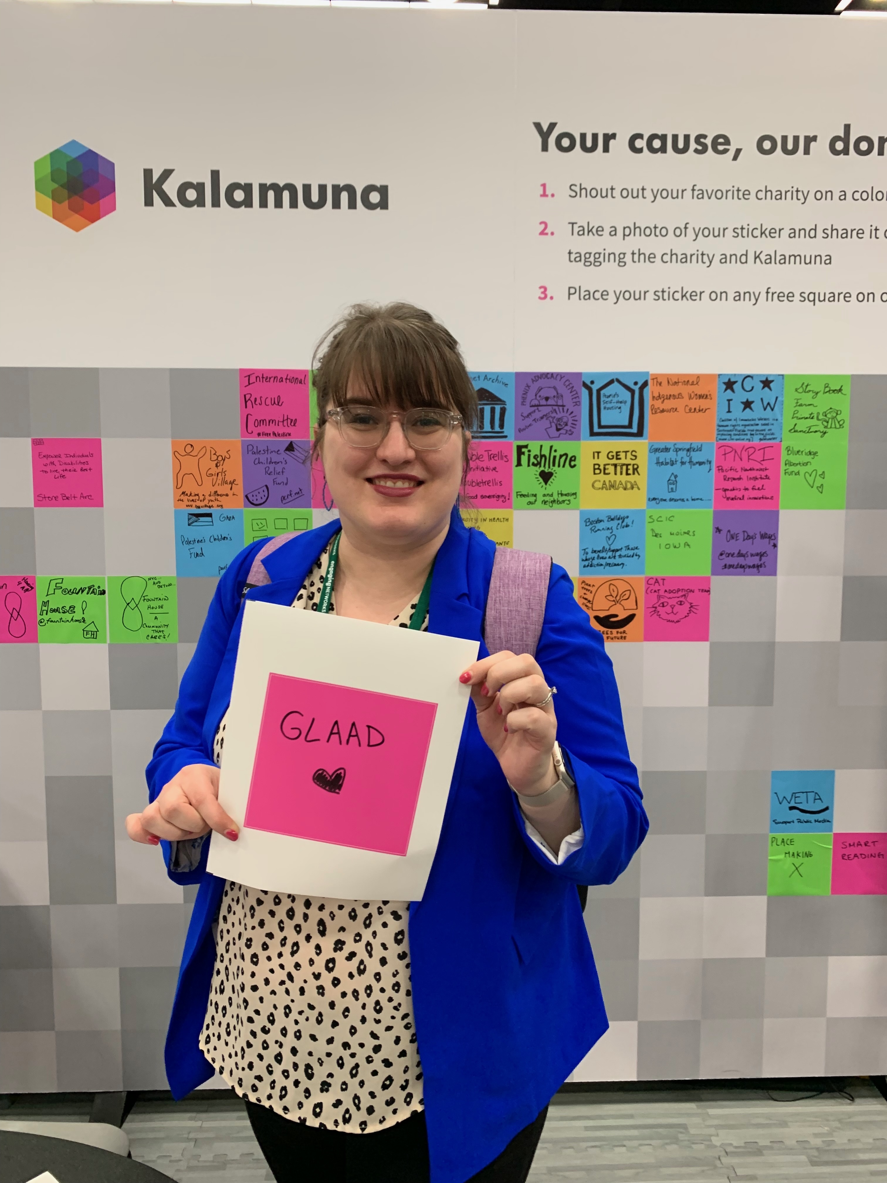 Emily Harden, a white women with clear glasses and straight across bangs, wearing a bright dark blue blazer and pattern shirt, holding a sign that says 'GLAAD' with a heart