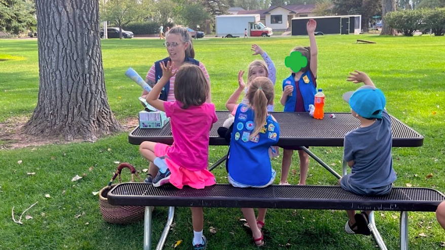 Jessica sitting with a group of Girl Scouts on a picnic table with all their hands in the air in a park with green grass.