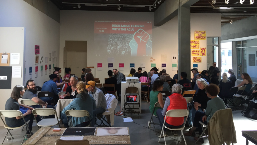 Image of a large room with many people sitting in chairs chatting with a projector with the words displaying "Resistance Trainer with the ACLU"