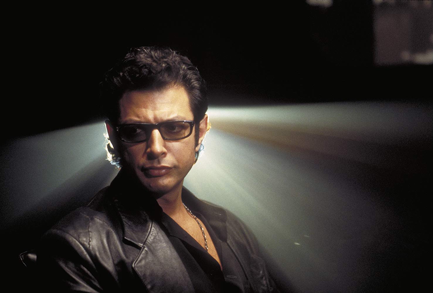 Jeff Goldblum looking serious in a leather jacket and sunglasses, silhouetted from behind.