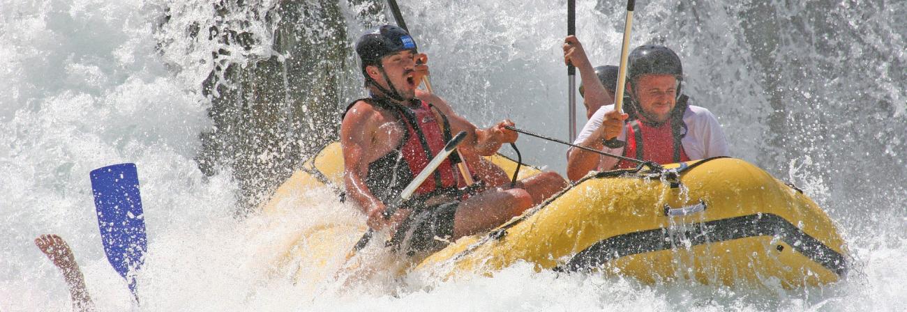 Photo of several people white water rafting and one person overboard with their paddle and foot in the air.