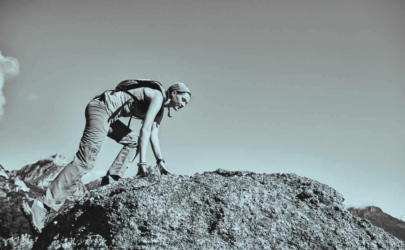 A woman climbing the tip of a mountain lunging onto the top with a blue greyish overtone on the image.