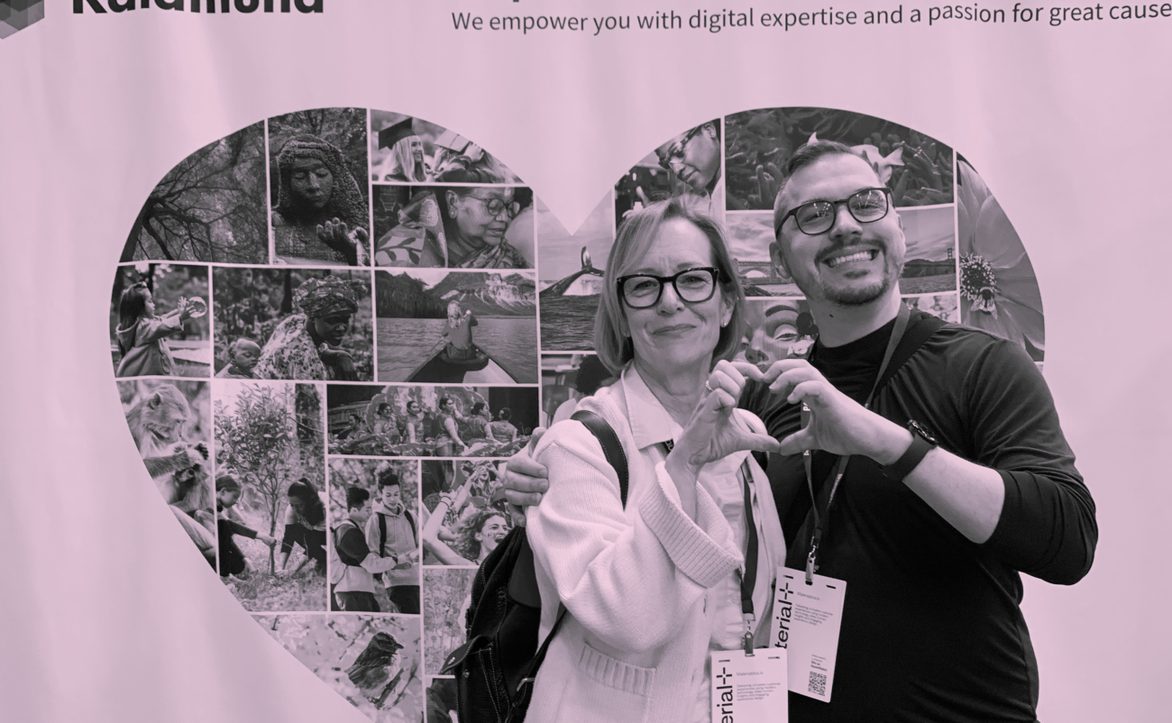 Two people making a heart with the hands in front of Kalamuna's conference booth that is a bunch of photos in the shape of a heart with a pink hue overlay filter on the photo. 