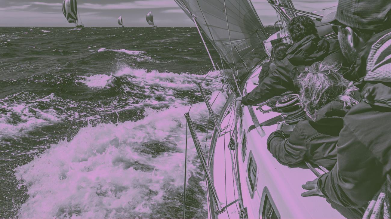Several people leaning along one edge of a sailboat in choppy waters, with several other sailboats in the distance 